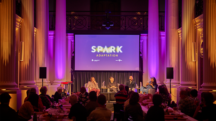 Audience seated around long dining tables at Spark: adaptation event, facing the stage where panelists are seated with a screen projecting the words 'Spark: adaptation' behind them