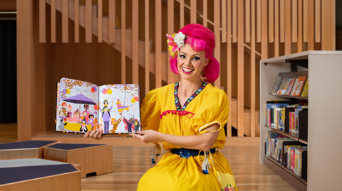A woman with pink hair, wearing a yellow dress, is seated, smiling, holding a picture book up.